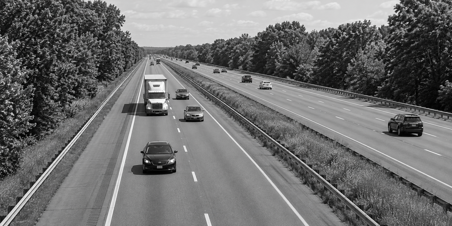 Black-and-white divided highway with a tractor-trailer and cars between tree-lined shoulders